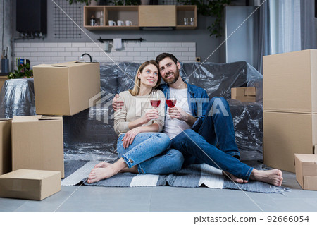 Family couple in love, man and woman sitting on the floor celebrating a new home in a new apartment, drinking wine among cardboard boxes 92666054