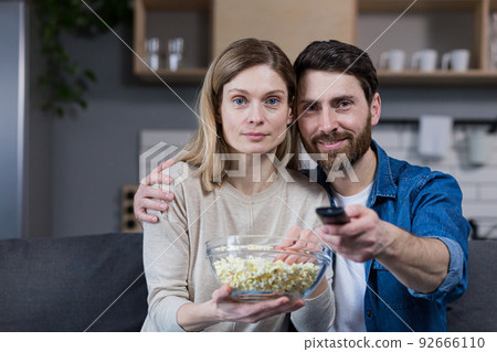 Photo of close couple man and woman together at home having fun and happily watching TV and eating popcorn Photo of close couple man and woman together at home having fun and happily watching TV and eating popcorn 92666110
