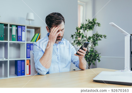 Upset and sad businessman looking at phone screen, man working in bright office Upset and sad businessman looking at phone screen, man working in bright office 92670163