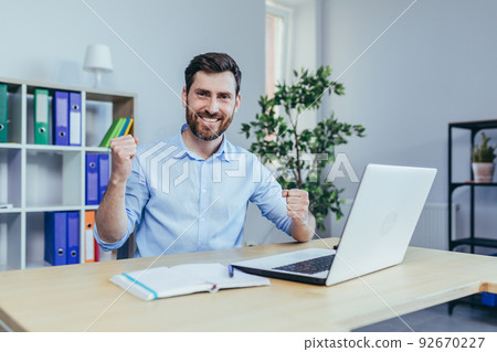 Successful businessman with a beard looks at the camera and smiles, celebrates triumph, works on a laptop in the office 92670227