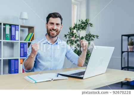 Successful businessman with a beard looks at the camera and smiles, celebrates triumph, works on a laptop in the office Successful businessman with a beard looks at the camera and smiles, celebrates triumph, works on a laptop in the office 92670228