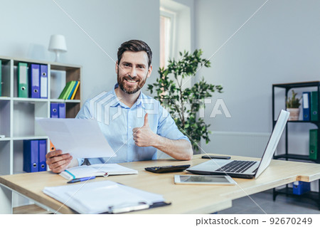 Successful businessman auditor looks at the camera and smiles, performs reconciliation of business reports, shows the camera a thumbs up Successful businessman auditor looks at the camera and smiles, performs reconciliation of business reports, shows the camera a thumbs up 92670249