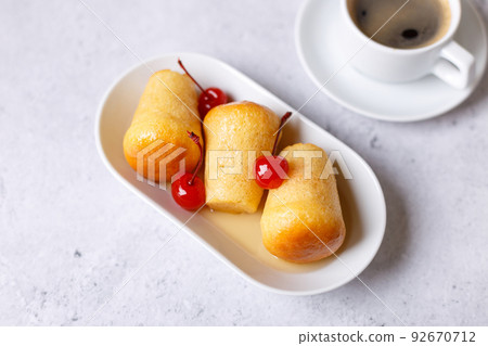 Neapolitan Rum baba (or baba au rum) on a white plate with a cocktail cherry and a cup of coffee. Small yeast cakes soaked in rum syrup on a gray background. Traditional Italian pastry. Close-up, sele Neapolitan Rum baba (or baba au rum) on a white plate with a cocktail cherry and a cup of coffee. Small yeast cakes soaked in rum syrup on a gray background. Traditional Italian pastry. Close-up, sele 92670712