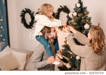 Happy family with child decorate christmas tree. Little girl sitting on daddy's shoulders hangs ball on the Christmas tree 92670729