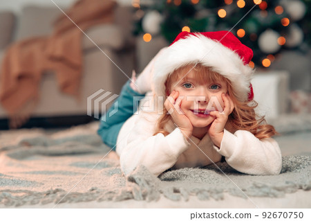 Beautiful little girl with white curly hair and Christmas hat lies in the room on the floor against the background of decorated Christmas tree and lights Beautiful little girl with white curly hair and Christmas hat lies in the room on the floor against the background of decorated Christmas tree and lights 92670750