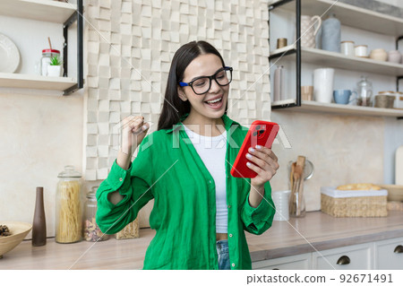 Beautiful young woman in glasses and green shirt read good news from red phone, brunette at home in kitchen shocked by good news 92671491