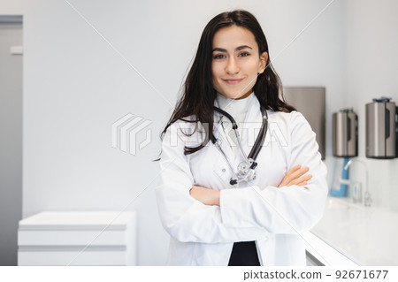 Portrait of cheerful female doctor in lab coat standing with crossed hands on her office Portrait of cheerful female doctor in lab coat standing with crossed hands on her office 92671677