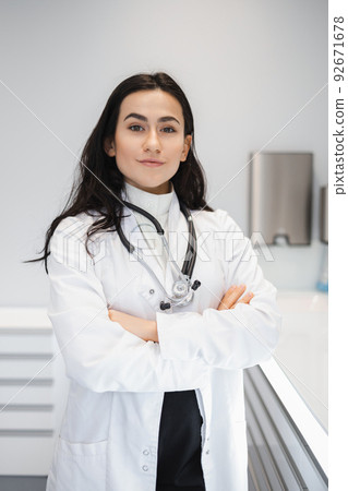 Portrait of female doctor in lab coat standing with crossed hands on her office 92671678