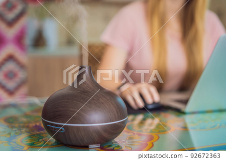 Essential oil diffuser on the table steaming while woman using laptop Essential oil diffuser on the table steaming while woman using laptop 92672363