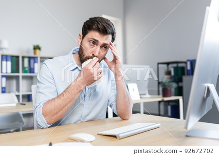 Sick and tired young man at work. Office worker, manager, freelancer sitting at a desk in the office does not feel well, wipes his nose, has a runny nose, coughs, headaches, holds his head. 92672702