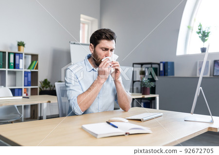 Businessman sick in the office man with allergies sneezes, sitting at a table with a computer Businessman sick in the office man with allergies sneezes, sitting at a table with a computer 92672705