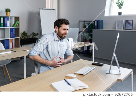 Office worker, freelancer, manager, young man feels abdominal pain, nausea, holding his stomach. Sitting at his desk at the computer, needs help Office worker, freelancer, manager, young man feels abdominal pain, nausea, holding his stomach. Sitting at his desk at the computer, needs help 92672706