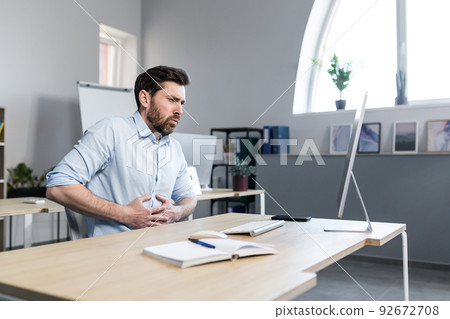 Office worker, freelancer, manager, young man feels abdominal pain, nausea, holding his stomach. Sitting at his desk at the computer, needs help Office worker, freelancer, manager, young man feels abdominal pain, nausea, holding his stomach. Sitting at his desk at the computer, needs help 92672708