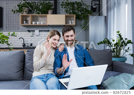 Young couple, family, man and woman sitting at home on the couch talking on video call from laptop to family, friends. They wave, smile, greet 92672825
