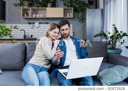Married couple man and woman sitting together at home on the couch using laptop for video chat and chatting with friends 92672826