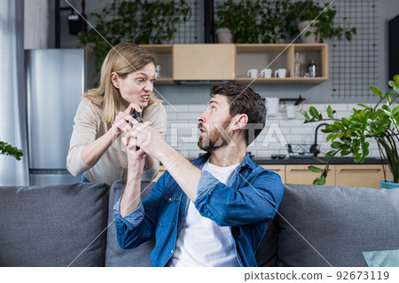 Conflict in the family. An angry, annoyed woman's wife takes the phone from the man who is sitting on the couch at home and constantly uses the phone. Conflict in the family. An angry, annoyed woman's wife takes the phone from the man who is sitting on the couch at home and constantly uses the phone. 92673119