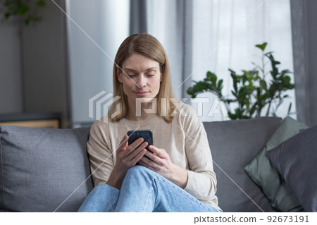 Beautiful middle-aged woman sitting on the couch at home, using the phone and smiling Beautiful middle-aged woman sitting on the couch at home, using the phone and smiling 92673191