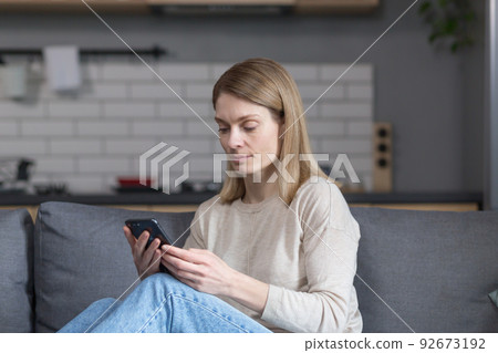 Beautiful middle-aged woman sitting on the couch at home, using the phone and smiling Beautiful middle-aged woman sitting on the couch at home, using the phone and smiling 92673192