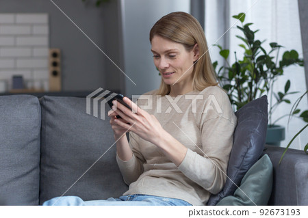 Beautiful middle-aged woman sitting on the couch at home, using the phone and smiling Beautiful middle-aged woman sitting on the couch at home, using the phone and smiling 92673193