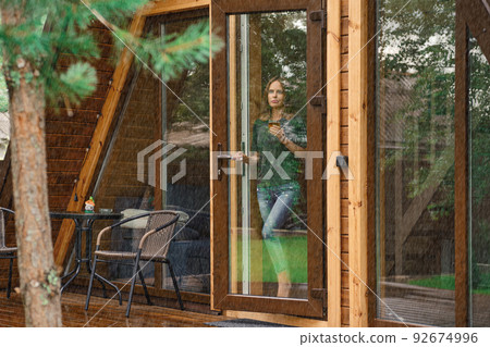 Young woman with a cup of herbal tea stands on the doorstep of a forest house and looks at the rain Young woman with a cup of herbal tea stands on the doorstep of a forest house and looks at the rain 92674996