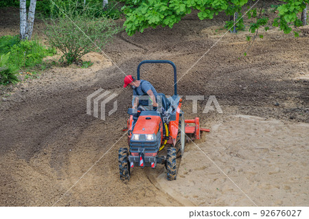A farmer on a mini tractor loosens the soil for the lawn. Land cultivation, surface leveling 92676027