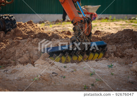 A mini excavator with a rake loosens the ground. landscape preparation. A mini excavator with a rake loosens the ground. landscape preparation. 92676047