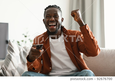 Overjoyed black man watching sports game on TV and celebrating victory of favorite team, sitting on sofa at home Overjoyed black man watching sports game on TV and celebrating victory of favorite team, sitting on sofa at home 92676700