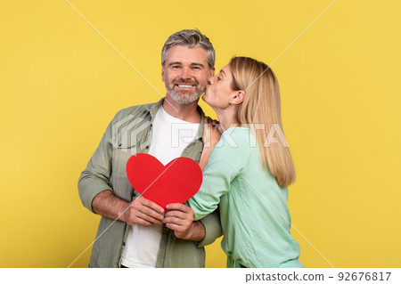 St. Valentines day concept. Loving mature spouses holding red paper heart card, woman kissing her happy husband St. Valentines day concept. Loving mature spouses holding red paper heart card, woman kissing her happy husband 92676817
