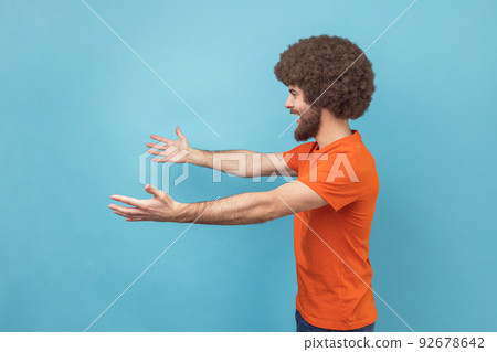 Come into my arms. Side view of satisfied man with Afro hairstyle wearing orange T-shirt reaching out to camera, stretching arms to hug you. Indoor studio shot isolated on blue background. Come into my arms. Side view of satisfied man with Afro hairstyle wearing orange T-shirt reaching out to camera, stretching arms to hug you. Indoor studio shot isolated on blue background. 92678642