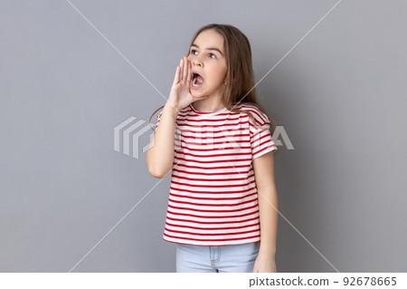 Attention, announcement. Portrait of dark haired adorable little girl wearing striped T-shirt screaming message, loud advertising. Indoor studio shot isolated on gray background. Attention, announcement. Portrait of dark haired adorable little girl wearing striped T-shirt screaming message, loud advertising. Indoor studio shot isolated on gray background. 92678665