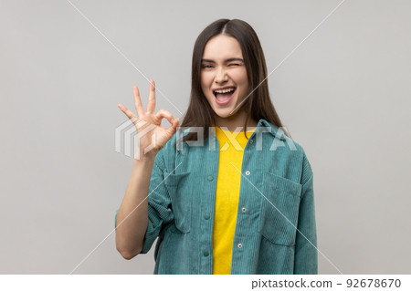 Portrait of satisfied beautiful young woman standing, looking at camera showing Ok sign gesture and winking, wearing casual style jacket. Indoor studio shot isolated on gray background. 92678670
