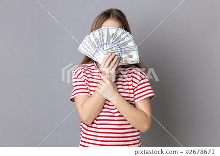 Portrait of unknown anonymous rich little girl wearing striped T-shirt hiding her face behind dollars banknotes, income of money. Indoor studio shot isolated on gray background. Portrait of unknown anonymous rich little girl wearing striped T-shirt hiding her face behind dollars banknotes, income of money. Indoor studio shot isolated on gray background. 92678671