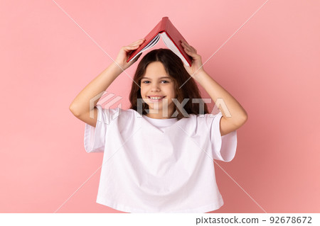 Portrait of smiling positive little girl wearing white T-shirt making roof, holding book over head, looking at camera with happiness. Indoor studio shot isolated on pink background. Portrait of smiling positive little girl wearing white T-shirt making roof, holding book over head, looking at camera with happiness. Indoor studio shot isolated on pink background. 92678672