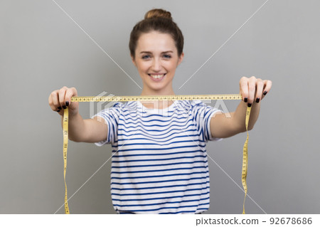 Portrait of joyful cheerful positive woman designer or seamstress wearing striped T-shirt showing measure tape, dressmaker working, measuring. Indoor studio shot isolated on gray background. 92678686