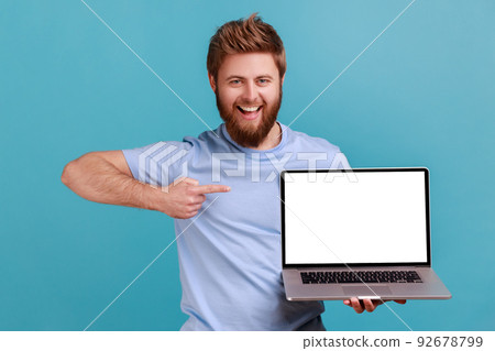 Portrait of happy bearded man holding laptop and pointing at empty screen, smiling satisfied with computer app, looking at camera. Indoor studio shot isolated on blue background. Portrait of happy bearded man holding laptop and pointing at empty screen, smiling satisfied with computer app, looking at camera. Indoor studio shot isolated on blue background. 92678799