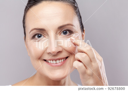 Closeup portrait of middle aged woman applies face cream, enjoys new anti wrinkle cosmetic product, prevents sign of skin aging, wears minimal makeup. Indoor studio shot isolated on gray background. Closeup portrait of middle aged woman applies face cream, enjoys new anti wrinkle cosmetic product, prevents sign of skin aging, wears minimal makeup. Indoor studio shot isolated on gray background. 92678800