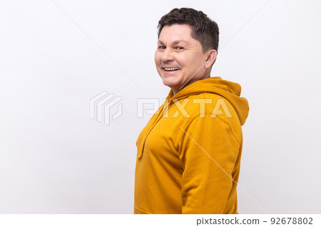 Side view of handsome middle aged man with dark hair standing looking at camera with satisfied face and smiling, wearing urban style hoodie. Indoor studio shot isolated on white background. Side view of handsome middle aged man with dark hair standing looking at camera with satisfied face and smiling, wearing urban style hoodie. Indoor studio shot isolated on white background. 92678802