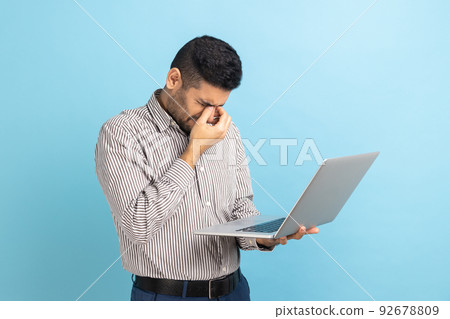 Portrait of young adult businessman standing with laptop in hands, rubbing his eyes, looks tired and exhausted, wearing striped shirt. Indoor studio shot isolated on blue background. 92678809