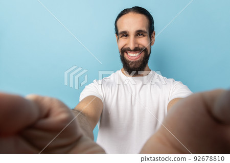 Man blogger with beard in white T-shirt looking at camera with toothy smile, being in good mood, broadcasting livestream, POV, point of view of photo. Indoor studio shot isolated on blue background. 92678810