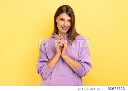 Portrait of devious cunning young woman clasping hands and smirking mysteriously, scheming cheats, evil prank, wearing purple hoodie. Indoor studio shot isolated on yellow background. 92678813