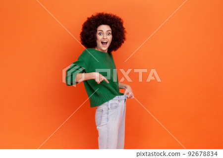 Happy woman with Afro hairstyle wearing green casual style sweater showing slim waist in big trousers, successful weight loss, diet conception. Indoor studio shot isolated on orange background. 92678834