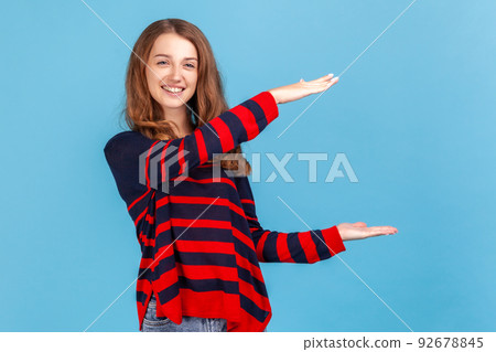Optimistic woman wearing striped casual style sweater, presenting copy space between hands, holding empty place for commercial text or image. Indoor studio shot isolated on blue background. 92678845