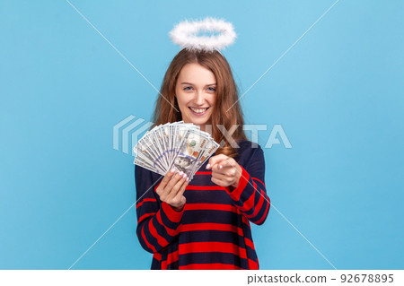 Angelic woman wearing striped casual style sweater and nimb over head, holding currency and pointing at camera, choosing you as a winner. Indoor studio shot isolated on blue background. Angelic woman wearing striped casual style sweater and nimb over head, holding currency and pointing at camera, choosing you as a winner. Indoor studio shot isolated on blue background. 92678895