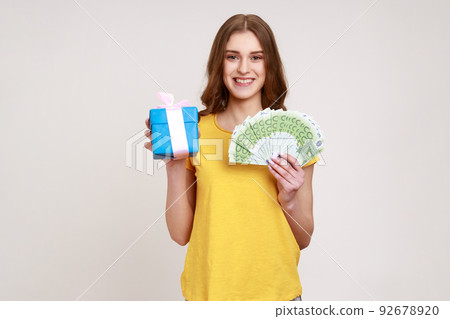 Gift shopping. Happy teenager girl in yellow T-shirt holding present box and euro money banknotes, satisfied with purchase, cashback and bank loan. Indoor studio shot isolated on gray background. 92678920