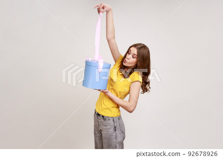 Portrait of surprised beautiful brown haired woman in yellow casual T-shirt, looking at ribbon on gift box with amazement, unboxing present. Indoor studio shot isolated on gray background. Portrait of surprised beautiful brown haired woman in yellow casual T-shirt, looking at ribbon on gift box with amazement, unboxing present. Indoor studio shot isolated on gray background. 92678926