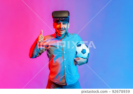 Portrait of man in shirt wearing virtual reality glasses, standing with soccer ball, showing thumb up, like football match. Indoor studio shot isolated on colorful neon light background. 92678939