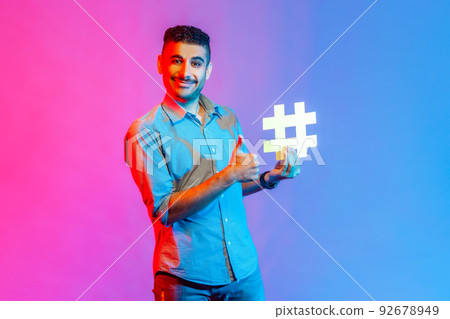 Portrait of smiling man in shirt holding hashtag symbol, showing thumb up, recommending popular topics, internet trends. Indoor studio shot isolated on colorful neon light background. Portrait of smiling man in shirt holding hashtag symbol, showing thumb up, recommending popular topics, internet trends. Indoor studio shot isolated on colorful neon light background. 92678949