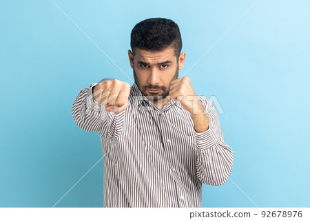 Portrait of aggressive young adult businessman standing with boxing fists and ready to attack or defence, looking with angry face, wearing striped shirt. Indoor studio shot isolated on blue background 92678976