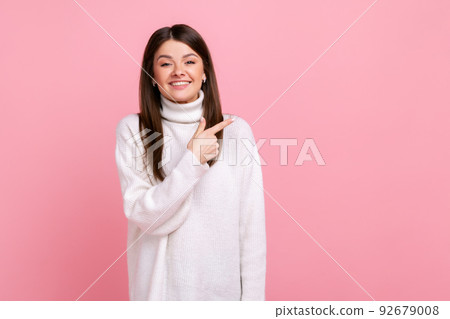 Charming young girl pointing aside at empty place for ad content, copy space for commercial product, wearing white casual style sweater. Indoor studio shot isolated on pink background. 92679008