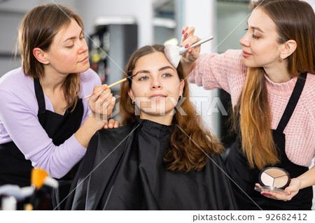 Two makeup artists apply makeup on the face of a young woman Two makeup artists apply makeup on the face of a young woman 92682412
